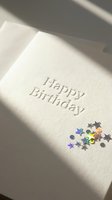 Elegant white Happy Birthday card with embossed text and holographic star confetti, lit by soft natural sunlight for a minimal celebration concept.