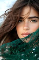Close-up portrait of young woman with green eyes and flowing brown hair, partially covered with emerald lace fabric, natural makeup and freckles on soft glowing skin.