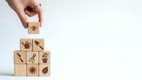 Hand placing wooden cube with gardening icons into pyramid arrangement on white background for spring planting and cultivation concepts.