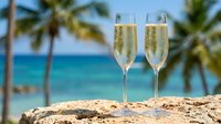 Two champagne flutes filled with sparkling wine on tropical beach with palm trees and turquoise ocean background for celebration and vacation concepts.