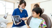 Asian female dentist in blue scrubs showing dental X-ray on tablet to young Asian boy patient in dental chair, explaining treatment in modern clinic.