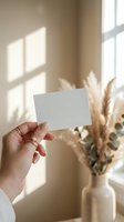 Female hand holding blank white card mockup in sunlit room with pampas grass and eucalyptus in ceramic vase for branding design presentation.