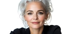 Elegant mature Caucasian woman with silver gray hair and natural makeup wearing black blazer smiling confidently against white background, beauty portrait.