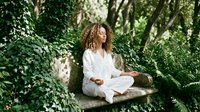 Young African American woman in white clothing practicing meditation in peaceful garden setting surrounded by lush green foliage and natural sunlight.