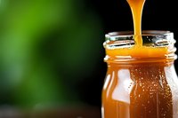 Golden honey dripping into glass jar with blurred green background, close-up macro shot showing viscous texture and amber color in natural lighting.