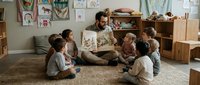 Male teacher with beard, Middle Eastern or Mediterranean ethnicity, reading a storybook to a diverse group of young children sitting on carpet in a bright classroom with artwork and wooden furniture.