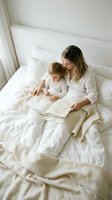 Young Caucasian mother and toddler son reading book together in white bed during cozy morning routine in bright bedroom for family bonding content.