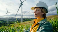 Young female engineer in safety helmet and yellow vest inspecting wind turbine farm on green hills against cloudy sky, renewable energy concept.