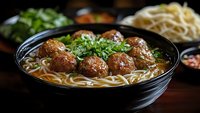 Asian meatball noodle soup in black bowl with fresh herbs and vegetables on wooden table for restaurant menu and food photography.