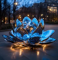 Glowing blue lotus flower sculpture illuminated at dusk with warm fairy lights, creating magical ambiance in urban park setting against bokeh city lights.