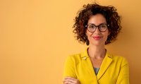 Confident woman with curly hair and glasses wearing bright yellow blazer against matching yellow background, arms crossed with friendly smile.