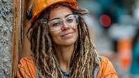 Young African American woman construction worker with dreadlocks wearing orange hard hat and safety glasses smiling confidently at camera on job site.