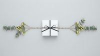 White gift box with black ribbon bow surrounded by eucalyptus branches and small flowers on light gray background, flat lay top view.
