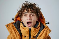 Young Caucasian male with curly brown hair wearing orange winter jacket shows excited surprised expression with open mouth against light background.