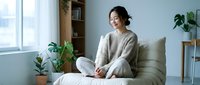 Young Asian woman in cozy sweater sitting peacefully on bed in bright modern bedroom with plants, enjoying quiet morning moment for wellness and relaxation.