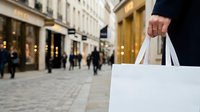 Shopper holding white paper bag on urban street with blurred storefronts and pedestrians in background, representing retail shopping and consumer lifestyle.