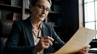 Mature woman professional wearing glasses and dark blazer reviewing documents at desk in modern office with bookshelves, suitable for business, corporate, and workplace concepts.