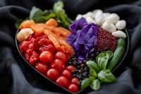 Fresh colorful vegetables and edible flowers arranged in heart shaped bowl with cherry tomatoes, carrots, mushrooms and purple pansies on dark background.