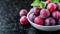 Fresh ripe plums covered with water drops in white ceramic bowl on dark background. Close up view of juicy purple fruits with morning dew.
