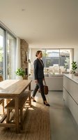 Woman in business attire walking through modern kitchen with wooden table, white cabinetry, and large windows overlooking living area. Professional home office setting.