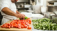 Middle Eastern male chef in white uniform precisely cuts fresh vegetables in commercial kitchen, preparing ingredients for restaurant service period.