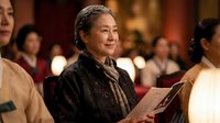 Mature Asian woman conductor leading orchestra performance in elegant concert hall with warm lighting and focused musicians in background.