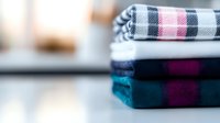 Neatly folded stack of plaid and checkered cotton towels in blue, purple, and gray tones, arranged on white surface with soft background blur.