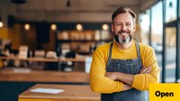 Confident mature male cafe owner in yellow sweater and denim apron standing with crossed arms in modern coffee shop interior, expressing positive business attitude.