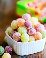 Colorful sugar-coated gummy candy balls in white ceramic bowl on wooden surface. Assorted fruit flavored sweets in pastel green, pink, and yellow colors.