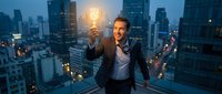 Young businessman holding glowing lightbulb on city rooftop at dusk representing innovation and creative business solutions for startup success.