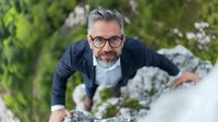 Mature businessman with gray hair and glasses climbing rocky terrain in nature, demonstrating determination and adventure in professional attire.