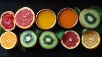 Fresh citrus fruits and kiwi slices arranged in row with orange and grapefruit juice glasses on dark background, healthy breakfast concept with green leaves.