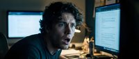 Middle-aged male with curly dark hair and Mediterranean ethnicity, wearing casual dark clothing, working late at computer workstation with multiple monitors displaying code or data. Stressed expression suggests deadline pressure or problem-solving in tech environment.