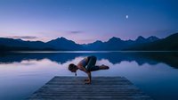 Young woman practicing crow pose yoga on wooden dock at serene mountain lake during twilight with crescent moon and purple sky for wellness meditation.
