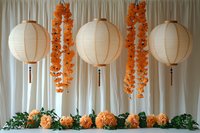 White paper lanterns with cascading orange floral garlands against white curtain backdrop, decorated with fresh flowers and greenery for wedding or celebration setup.