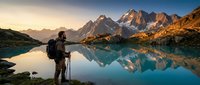 Male hiker with backpack and trekking poles admiring mountain lake reflection at golden hour sunrise. Adventure travel and outdoor recreation concept.