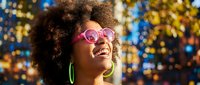 Happy African American woman with curly afro hair wearing pink sunglasses and green earrings smiling against colorful bokeh lights background for party celebrations.