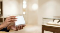 Woman's hand holding blank white card in luxury jewelry store with display cases and soft bokeh lighting in background.