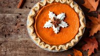 Fresh homemade pumpkin pie with whipped cream star decoration and anise on top, flaky crust edges, rustic wooden background with autumn maple leaves.