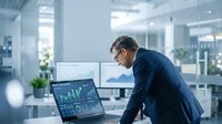 Middle-aged Caucasian businessman analyzing financial data charts on laptop in modern office workspace with glass partitions and natural lighting.