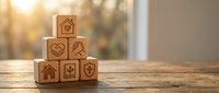Wooden blocks stacked in pyramid shape with engraved icons representing home, heart, handshake, care, growth, and protection on warm bokeh background.