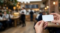 Close-up of hands holding blank white business card in busy coffee shop or cafe with blurred customers and warm lighting in background.
