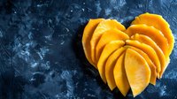 Fresh mango slices arranged in heart shape on dark blue textured background. Tropical fruit composition for healthy eating and romance concept.