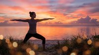 Young woman practicing yoga warrior pose at sunset beach with dramatic orange pink sky reflecting on calm ocean water creating peaceful meditation atmosphere.