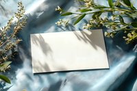 Blank white card mockup with natural shadow overlay from plants on blue silk fabric background, creating elegant composition for wedding invitations.