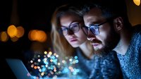 Young diverse couple looking at laptop screen in dark room with bokeh lights background, faces illuminated by blue screen glow, working late concept.
