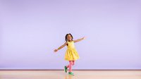 Young girl with dark skin tone wearing bright yellow dress and colorful sneakers, arms outstretched in joyful pose against soft purple background, expressing happiness and freedom.