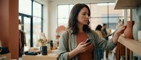Young woman shopping for shoes in modern retail store, examining footwear display while holding smartphone, casual lifestyle and consumer behavior concept.