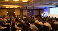 Large audience attending business conference presentation in modern hotel ballroom with professional speaker on stage and projection screen.