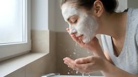 Woman with clay mask on face rinsing skin with water at bathroom sink during skincare routine and beauty treatment.